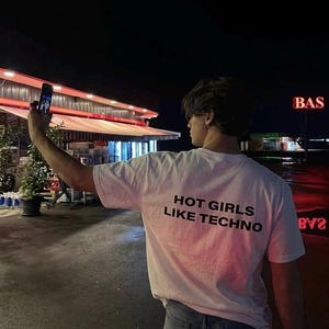 Può includere: Una maglietta bianca con la scritta "HOT GIRLS LIKE TECHNO" in nero. La persona indossa jeans blu e tiene in mano un telefono. Lo sfondo include un edificio con una tenda rossa e un'insegna con la scritta "BAS".