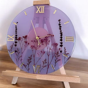 May include: A purple resin clock with dried flowers embedded in the resin. The clock face has Roman numerals and is set on a wooden easel.
