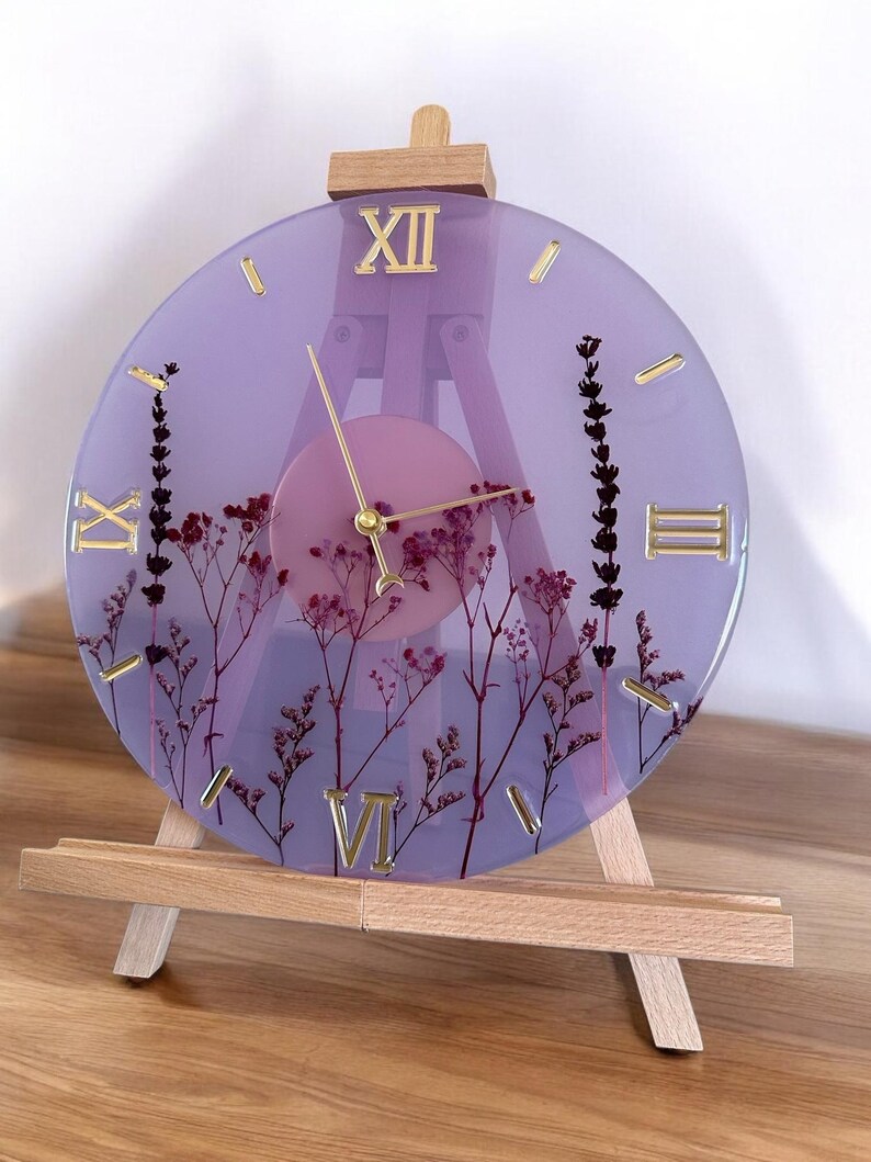 May include: A purple resin clock with dried flowers embedded in the resin. The clock face has Roman numerals and is set on a wooden easel.