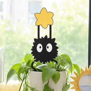 May include: A black soot sprite plant trellis with a yellow star topper, featuring two large white eyes and a spiky silhouette, inserted into a white ceramic pot containing a green leafy plant with trailing vines and stems.