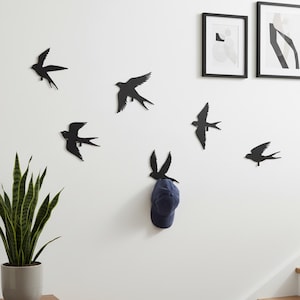 May include: Black metal swallow wall art decor, arranged in a flock-like pattern on a white wall. A navy blue baseball cap hangs from one of the swallows. The scene includes a potted plant and framed artwork.