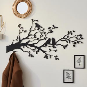 May include: A black metal wall art piece depicting a tree branch with leaves and three birds perched on it. The art piece is mounted on a white wall, with a round woven mirror and framed botanical prints nearby. A brown coat hangs below.