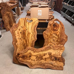 Op de afbeelding: Drie natuurlijke houten snijplanken met onregelmatige vormen en unieke houtnerfpatronen. De planken zijn gemaakt van olijfhout en hebben een lichtbruine kleur met donkerbruine strepen. De planken zijn perfect om kaas, charcuterie of andere hapjes op te serveren.