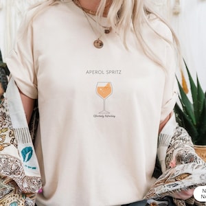 Puede incluir: Camiseta color crema con las palabras "APEROL SPRITZ" sobre un gráfico de un cóctel Aperol Spritz. El texto "Effortlessly Refreshing" está debajo de la imagen del cóctel. Parte de una colección de ropa.
