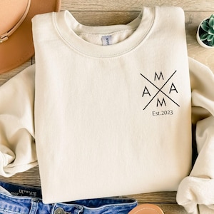 Könnte beinhalten: Cremefarbenes Sweatshirt mit dem Wort "MAMA" in einem durchgestrichenen Design, darunter "Est. 2023". Das Sweatshirt liegt flach mit einem braunen Hut und blauen Jeans im Bild. Eine kleine Topfpflanze ist ebenfalls zu sehen.