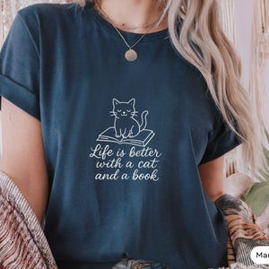 Könnte beinhalten: Marineblaues T-Shirt mit weißer Grafik einer Katze auf einem Buch. Darunter steht "Life is better with a cat and a book". Das Shirt hat einen lässigen Stil.