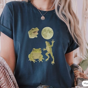 Può includere: Una t-shirt blu navy con una grafica di tre rane e una luna. Le rane sono verdi e hanno pose diverse. La luna è bianca e ha una superficie testurizzata.