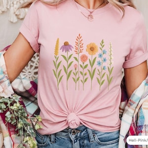 Può includere: Una maglietta rosa con un design floreale che presenta una varietà di fiori in tonalità di rosa, viola, giallo e arancione. I fiori sono disposti in una fila attraverso la parte anteriore della maglietta.