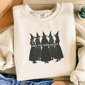 Può includere: Felpa color crema con grafica di cinque streghe con abiti e cappelli neri. Il testo "HEX THE PATRIARCHY" è stampato sull'immagine. La felpa è appoggiata su una superficie di legno, con un cappello e jeans visibili.