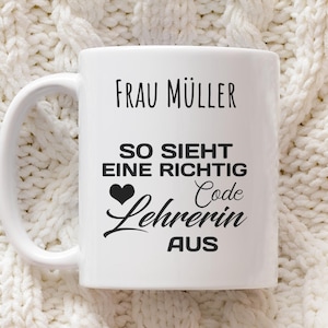 Könnte beinhalten: Weiße Keramiktasse mit Henkel, mit schwarzem Text, der "Frau Müller" oben liest, gefolgt von einem deutschen Satz und dem Wort "Code Lehrerin" mit einem Herzsymbol. Die Tasse steht vor einem weißen Strickhintergrund.
