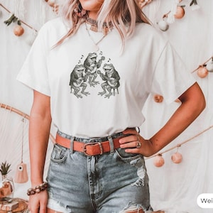 Può includere: Maglietta bianca con una grafica di tre rane che suonano strumenti musicali. Le rane indossano cappelli e tengono chitarre e un banjo.
