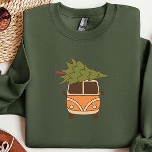 Könnte beinhalten: Ein olivgrünes Sweatshirt mit einer Cartoon-Illustration eines orangefarbenen und braunen Vans, der einen grünen Weihnachtsbaum auf dem Dach trägt. Der Van hat braune Räder und eine weiße Front. Das Sweatshirt ist ein Rundhals-Stil.