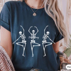 Op de afbeelding: Marineblauwe t-shirt met drie witte skeletfiguren in balletposes en de tekst "Stay Strong". Een casual t-shirt, ideaal voor dagelijks gebruik.