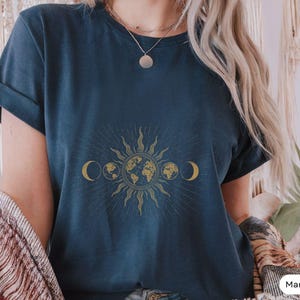 Op de afbeelding: Een marineblauw t-shirt met een gouden zon-, maan- en aarde-afbeelding. De afbeelding toont een gestileerde zon met stralen, een halve maan aan elke kant en een wereldbol in het midden.