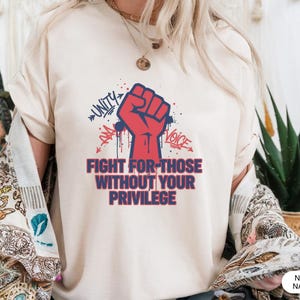 Może przedstawiać: Kremowa koszulka z czerwoną grafiką pięści i napisami "UNITY", "VOICE" oraz "FIGHT FOR THOSE WITHOUT YOUR PRIVILEGE" w postarzonej czcionce. Projekt zawiera niebieskie i czerwone akcenty.