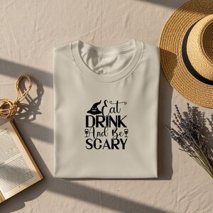 May include: A cream-coloured t-shirt featuring the black text "Eat Drink And Be Scary". The design includes a witch's hat, wine glasses, and a spooky face. A straw hat, a book, and lavender sprigs are also visible.
