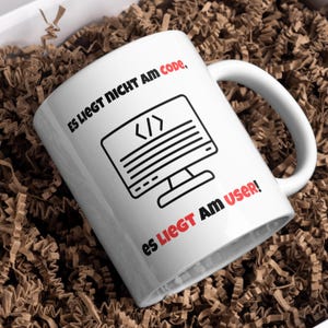 Op de afbeelding: Witte keramische mok met zwarte tekst die "Es liegt nicht am Code, es liegt am User!" luidt en een zwart-witte illustratie van een computerscherm met code op het scherm.