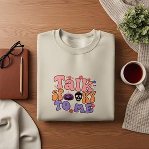 May include: Cream-colored sweatshirt with the phrase "Talk Spooky To Me" in a retro font. The design includes a purple pumpkin, a black skull, and flowers. A fun, festive top for Halloween.