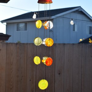 May include: A colorful wind chime made from glass and metal. The chime features a variety of round, yellow, orange, and clear glass pieces, as well as a red metal top.