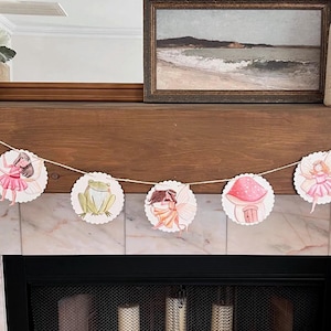 May include: A white and pink fairy themed banner with a snail, frog, mushroom, and ladybug. The banner is strung on a white string and is hanging above a fireplace.