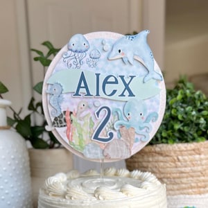 May include: Under the Sea cake topper with a blue and white dolphin, a jellyfish, an octopus, and a seahorse. The topper is decorated with a green banner that says "Alex 2".