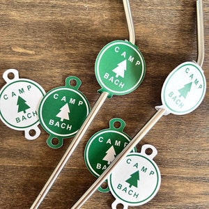 May include: Two silver metal reusable straws with green and white circular stickers that say "Camp Bach" with a green pine tree icon.