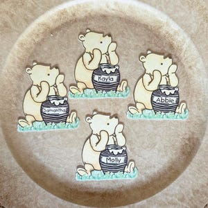 May include: Four wooden Winnie the Pooh-themed cutouts, each with a name on a honey pot. The bears are light brown, sitting on green grass, and the honey pots are black and white. Names include Kayla, Samantha, Abbie, and Molly.