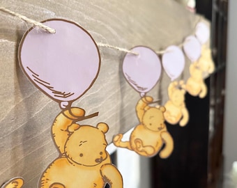 Pode incluir: Uma grinalda decorativa com uma série de recortes de um urso amarelo segurando um balão roxo claro. Cada urso é ilustrado numa posição sentada e suspenso por um fio fino de cor natural, criando um padrão repetitivo ao longo da corda.