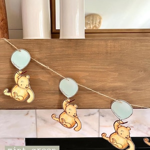 May include: A garland with three Winnie the Pooh characters holding light blue balloons. The garland is made of twine and the characters are printed on cardstock. The background is a wooden mantel with a white tile surround.