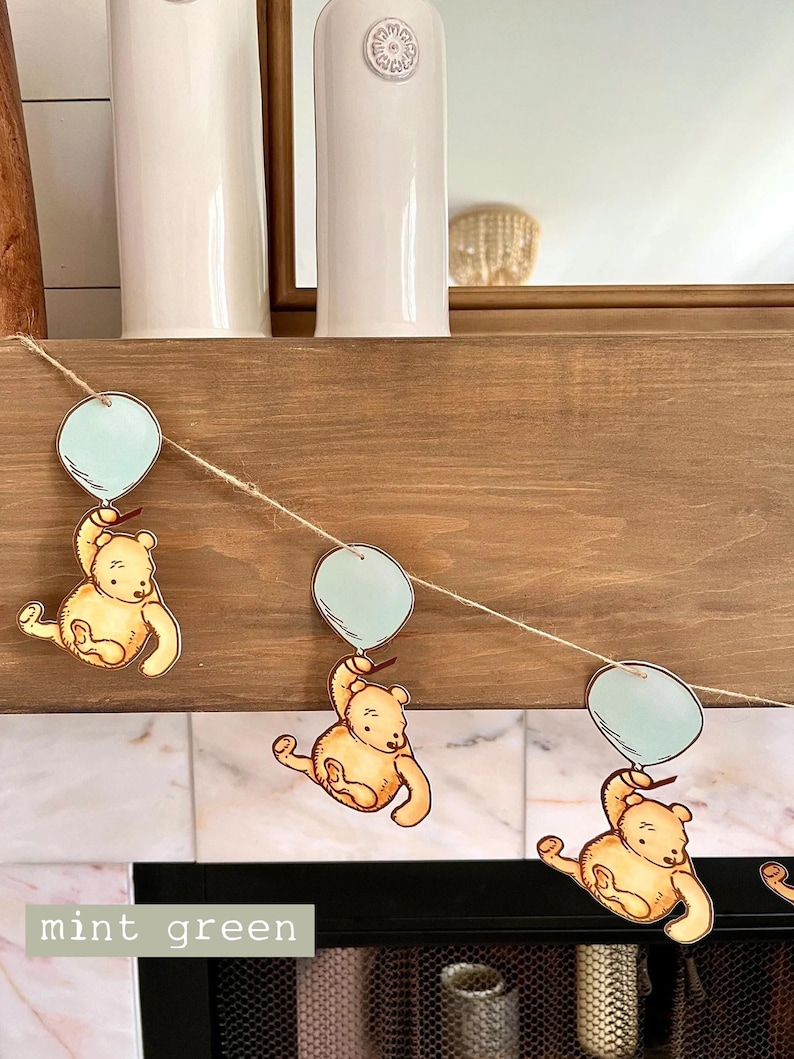 May include: A garland with three Winnie the Pooh characters holding light blue balloons. The garland is made of twine and the characters are printed on cardstock. The background is a wooden mantel with a white tile surround.