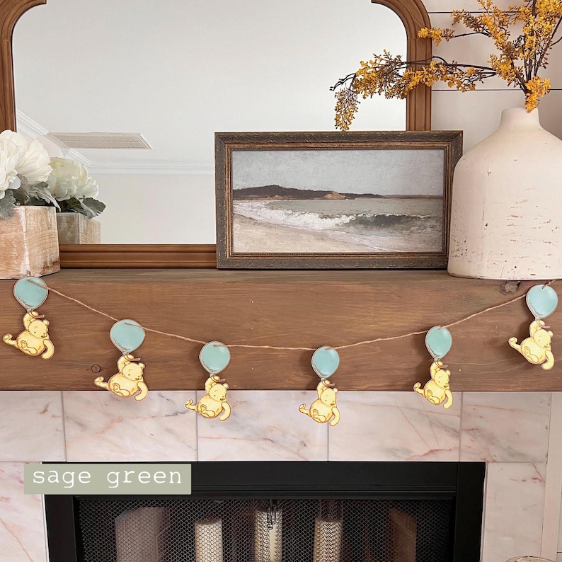 May include: A garland of Winnie the Pooh characters holding blue balloons. The garland is strung across a wooden mantel with a framed picture of a seascape above it. The text "sage green" is visible in the bottom left corner of the image.