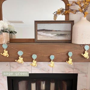 May include: A garland with a string of light blue balloons and yellow bear characters holding the balloons. The garland is hanging above a fireplace with a sage green mantle.