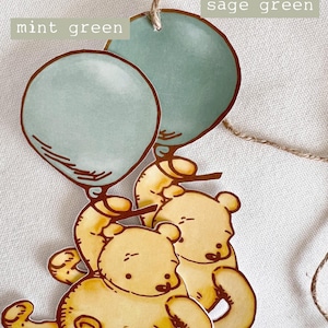 May include: Two yellow cartoon bears holding sage green and mint green balloons. The balloons are tied together with twine.