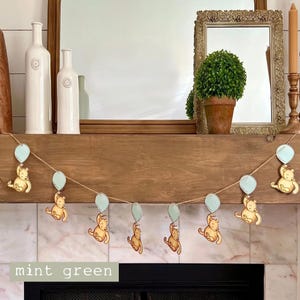 May include: A garland with a string of light blue balloons and Winnie the Pooh bears holding the balloons. The garland is hanging on a wooden mantel above a fireplace. The text "mint green" is written on the bottom of the image.