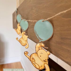 May include: A Winnie the Pooh themed baby shower banner with a string of yellow bears holding blue balloons. The banner is hanging on a wooden surface.