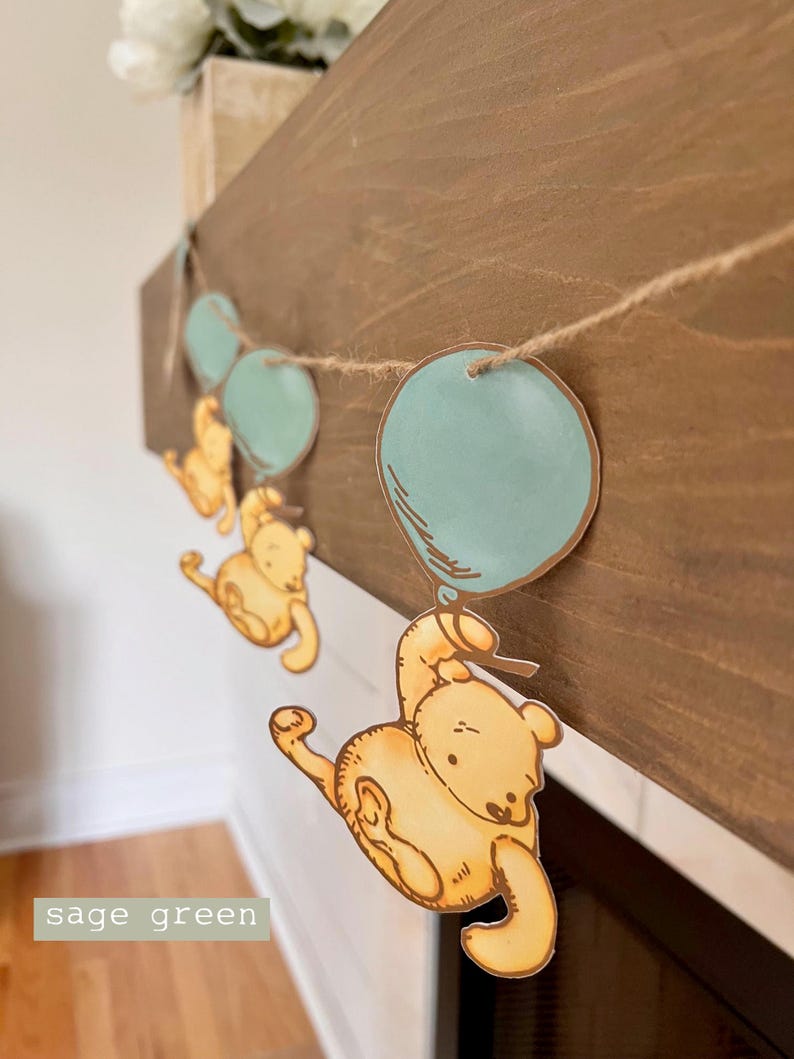 May include: A Winnie the Pooh themed baby shower banner with a string of yellow bears holding blue balloons. The banner is hanging on a wooden surface.