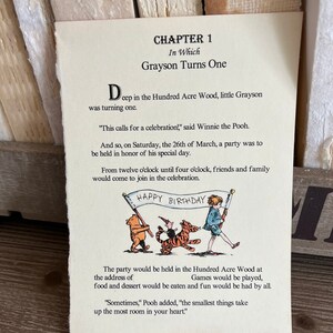 以下が含まれることがあります： 子供向けの本からの、くまのプーさんのイラスト。イラストには、くまのプーさん、ピグレット、ティガーが「ハッピーバースデー」と書かれた横断幕を持っています。ページのテキストには「第1章 グレイソンが1歳になるまで」と書いてあります。