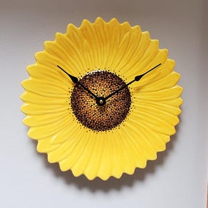May include: A decorative wall clock shaped like a sunflower. The clock face is a vibrant yellow with a textured, brown center. Black clock hands indicate the time. The clock is mounted on a light-colored wall.
