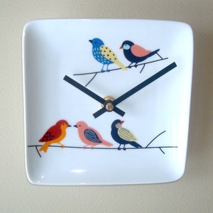 May include: A white ceramic square clock with black hands and a gold center. The clock face features a design of five colorful birds perched on a branch.