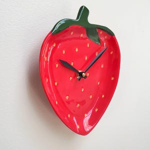 May include: A red strawberry-shaped wall clock with a green stem and leaves. The clock face features black hands and yellow seed details. The clock is mounted on a white wall.