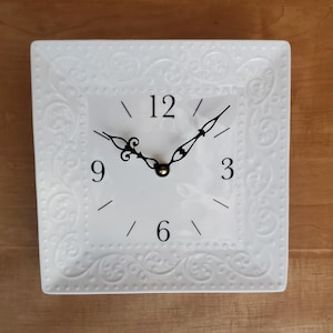 May include: White square clock with black Roman numeral markings and black hands. The clock face has a decorative white border with a raised design.