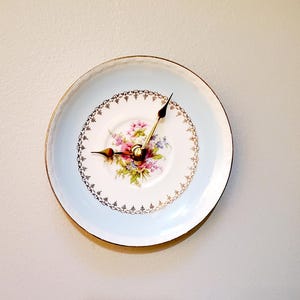 May include: A decorative wall clock made from a vintage plate. The plate features a light blue border and a floral center with pink, purple, and green flowers. Gold-colored clock hands and a gold rim complete the design.
