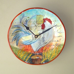 May include: A round, ceramic wall clock with a rooster design. The rooster is white with a red comb and wattle, and has blue and white feathers. The clock face is surrounded by a red border and has a black clock hand. The background is a watercolor-style design with wheat stalks and handwritten French words.