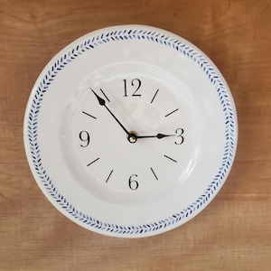 May include: White ceramic clock with blue and white patterned border. The clock face has black hands and black numbers.