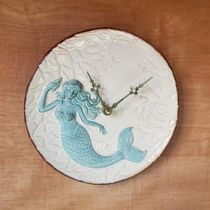 May include: A white ceramic clock with a blue mermaid design. The mermaid is depicted with long flowing hair and a tail. The clock face is surrounded by a white coral design.