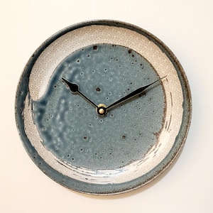 May include: A round ceramic clock with a blue-grey center and a patterned white border. The clock hands are black with a gold-colored center. The clock has a rustic, handmade appearance.