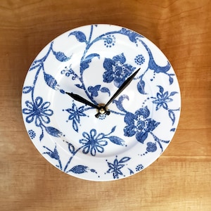 May include: White ceramic plate repurposed as a clock, featuring a blue floral design. The clock has black hands and a gold-colored center. The design includes stylized flowers and leaves, creating a decorative wall clock.