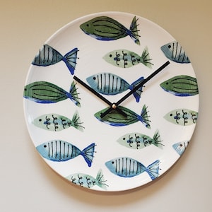 May include: A white ceramic clock with black hands and a blue and green fish design. The clock face is decorated with stylized fish in various shades of blue and green.