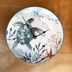May include: Round wall clock with a sea turtle design. The clock face features a watercolor illustration of a green sea turtle, coral, and fish against a white background. Black clock hands and a gold center complete the design. The clock has a green border.