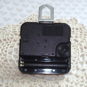 May include: A black quartz clock movement with a metal hanger. The clock mechanism has a battery compartment holding two AA batteries. The clock movement is on a cream-colored, crocheted doily.
